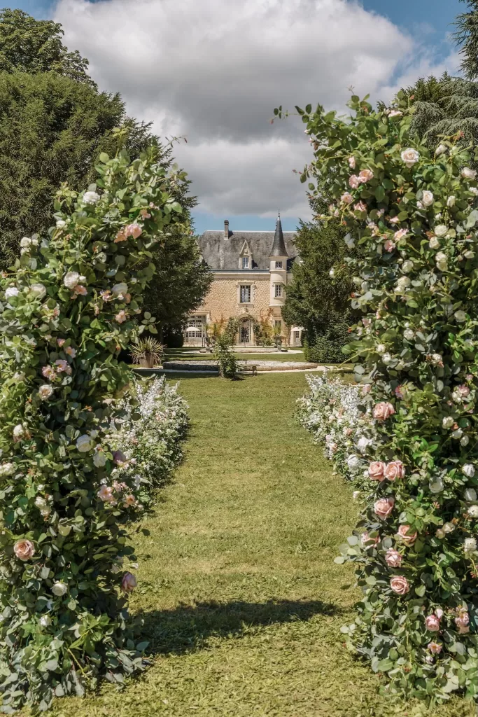 Romantic view through rose-covered ceremony arches towards Château de la Couronne’s fairytale facade.