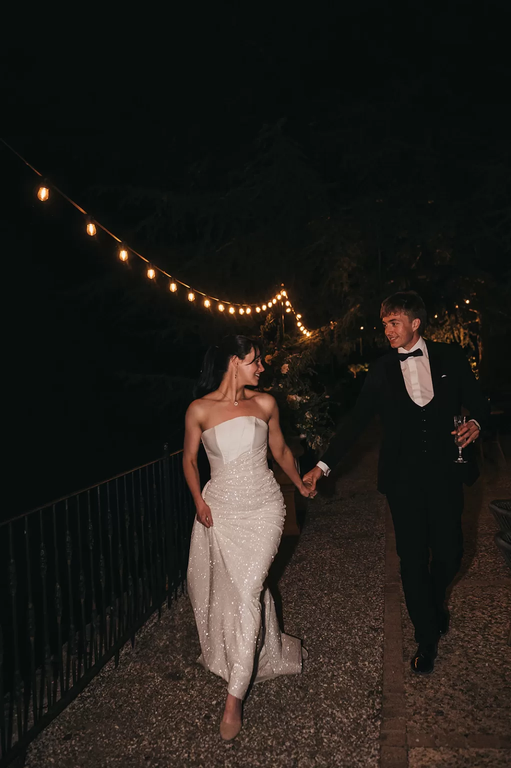Bride and groom walking hand in hand under string lights at night at Château de Sanse