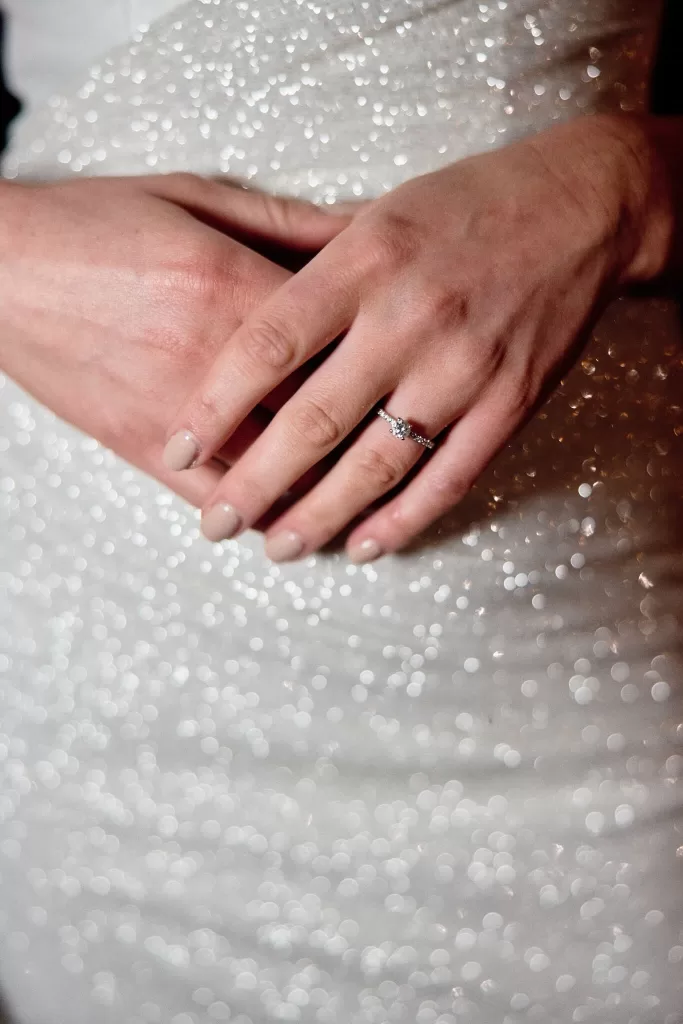 Close-up of engagement ring on bride’s hand against sparkling beaded gown.
