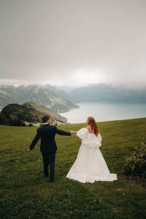 Bride and groom walk across an alpine meadow holding hands, with misty lake and mountain views below Hotel Villa Honegg.