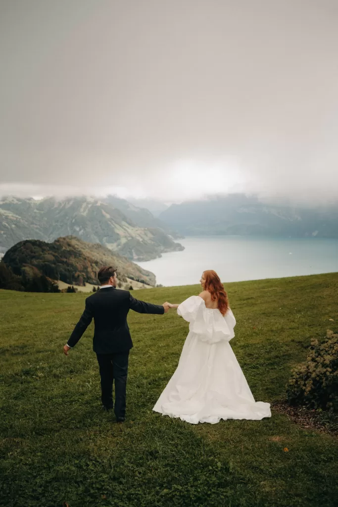 Bride and groom walk across an alpine meadow holding hands, with misty lake and mountain views below Hotel Villa Honegg.