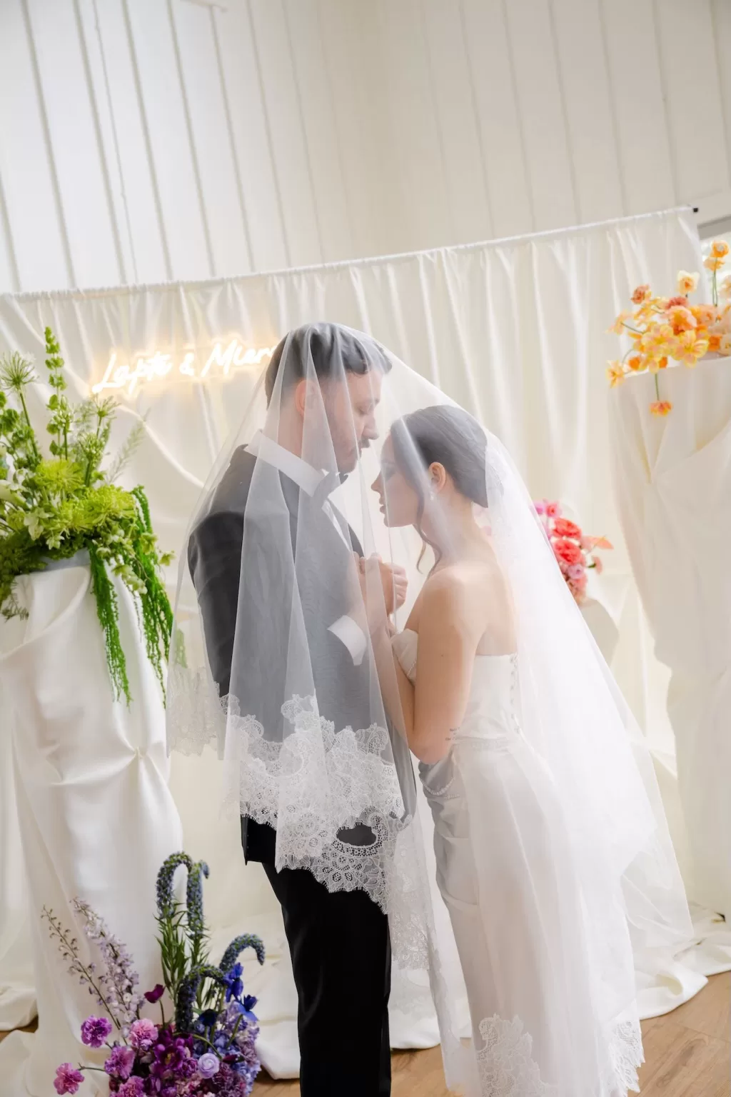 Intimate moment of bride and groom under lace veil during modern indoor ceremony.