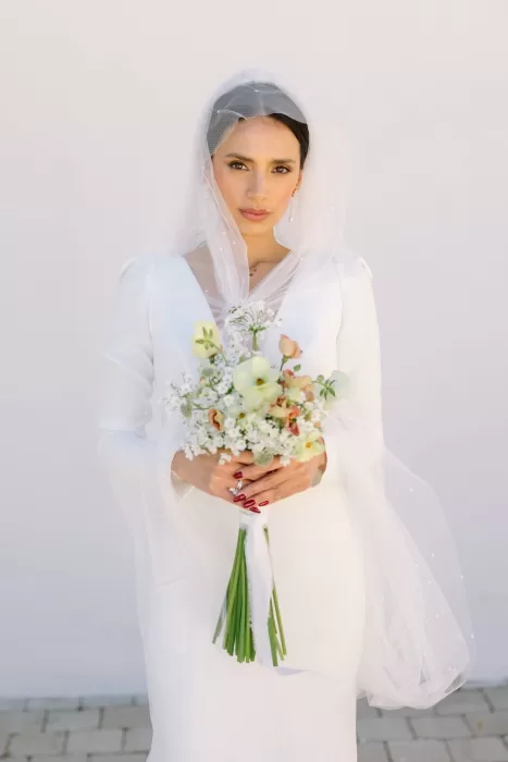 Bride in long-sleeved gown holding delicate bouquet against a sunlit white wall at Masia Cabellut