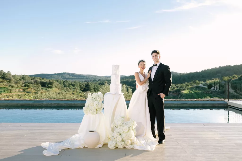 Bride and groom posing by the pool with elegant cake and floral décor at Masia Cabellut
