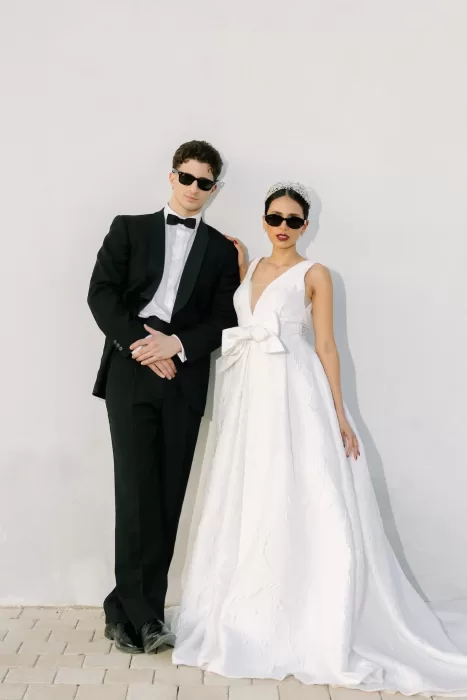 Stylish wedding couple in sunglasses leaning against white wall at Masia Cabellut