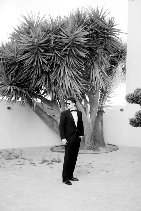 Groom in tuxedo standing beneath dramatic tropical foliage in the courtyard of Masia Cabellut.