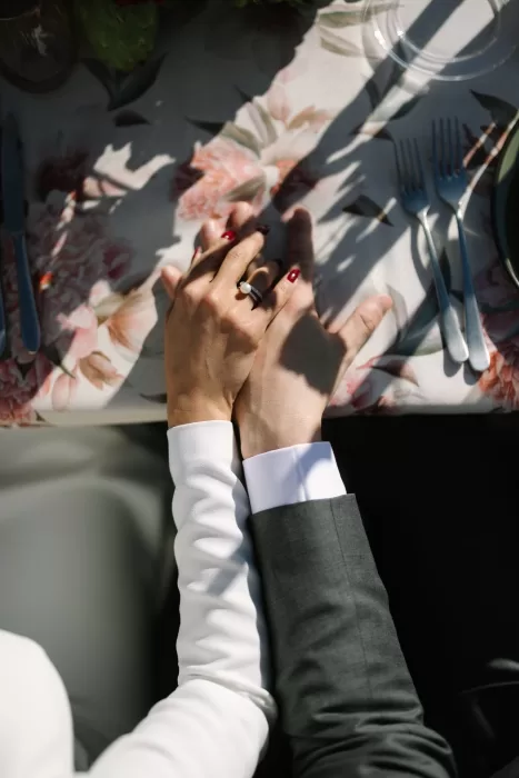 Close-up of bride and groom holding hands on floral tablecloth, wedding rings in focus.
