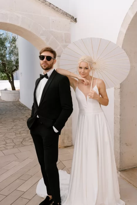 Stylish bride and groom standing beneath stone arches at Masseria San Giovanni, with the bride holding a paper parasol.