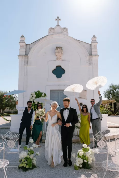 Joyful newlyweds exit the ceremony at Masseria San Giovanni, surrounded by cheering guests holding paper parasols.