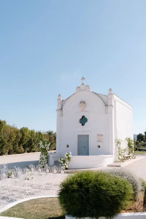 Outdoor wedding ceremony setup in front of the Masseria San Giovanni chapel, with white chairs and lush greenery.