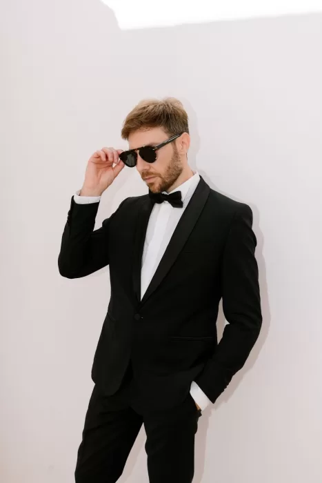 Groom in tuxedo adjusting sunglasses against sunlit white wall at Masseria San Giovanni