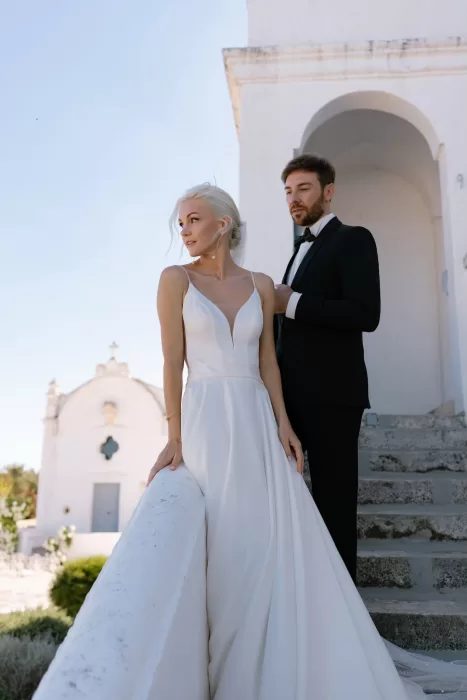 Bride and groom pose on the chapel steps of Masseria San Giovanni, surrounded by crisp white architecture and sunlight.