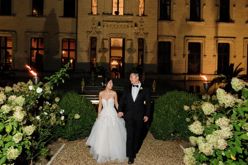 Bride and groom walking hand in hand through Château Challain’s torch-lit garden at night, smiling at each other.