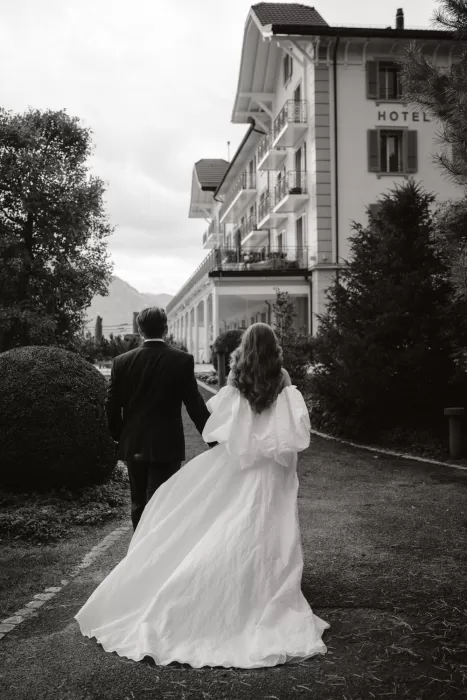Bride and groom walk hand in hand away from Hotel Villa Honegg, their elegant attire trailing behind.