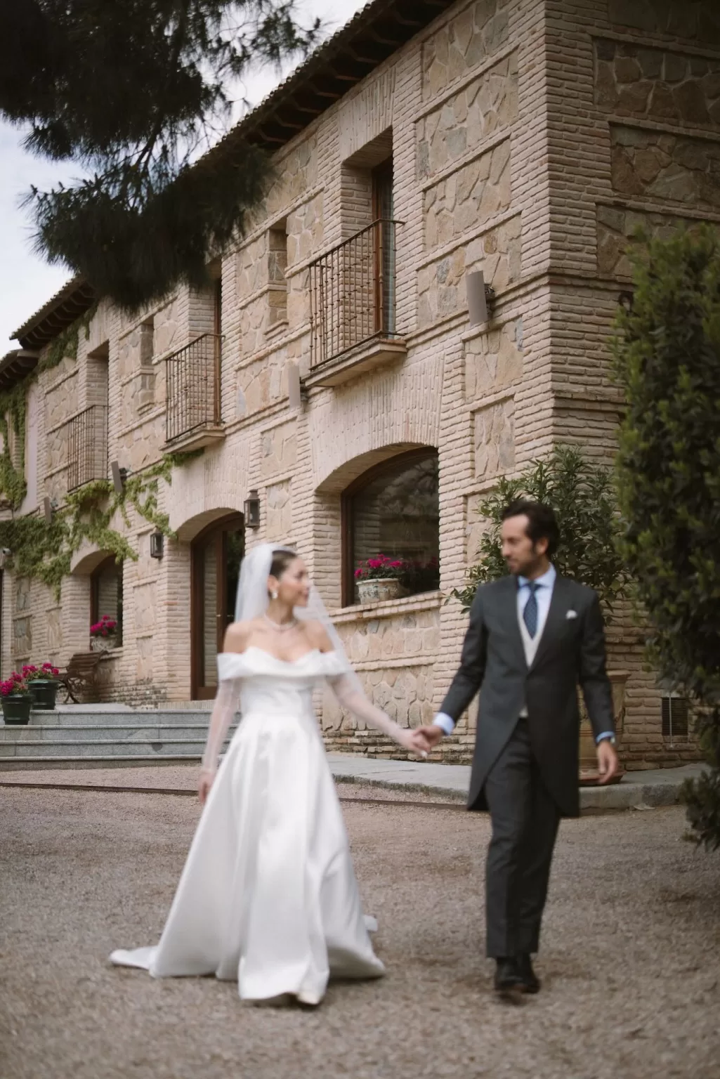 Bride and groom walk hand in hand in front of the rustic stone facade of Cigarral de la Mercedes.
