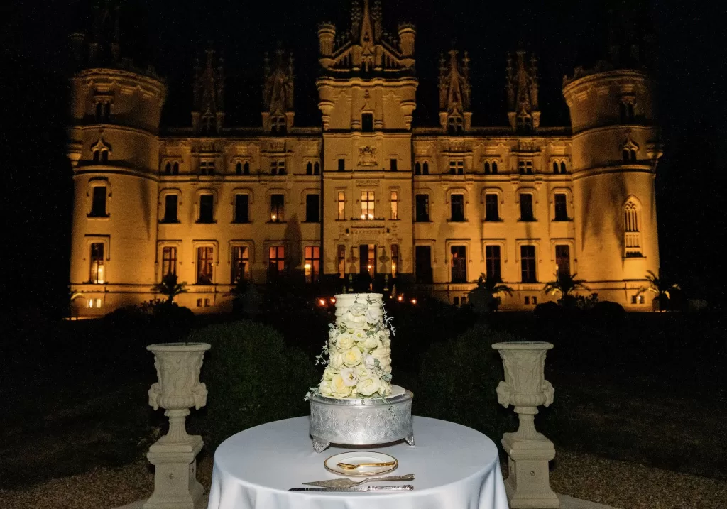 Tall white rose wedding cake glowing in front of the illuminated Château Challain at night.