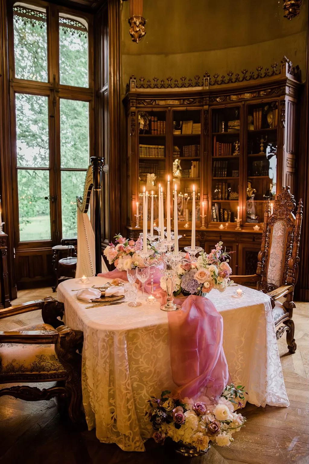 An opulent candlelit wedding dinner setup with pink draping and pastel florals in a grand wood-paneled library, photographed by Wim Vanhengel.