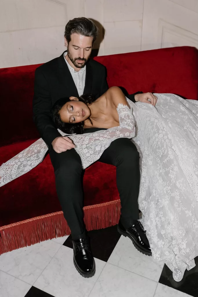 Bride rests peacefully in groom’s lap on a deep red velvet sofa, her lace gown draped around them, by Bahr Studios.