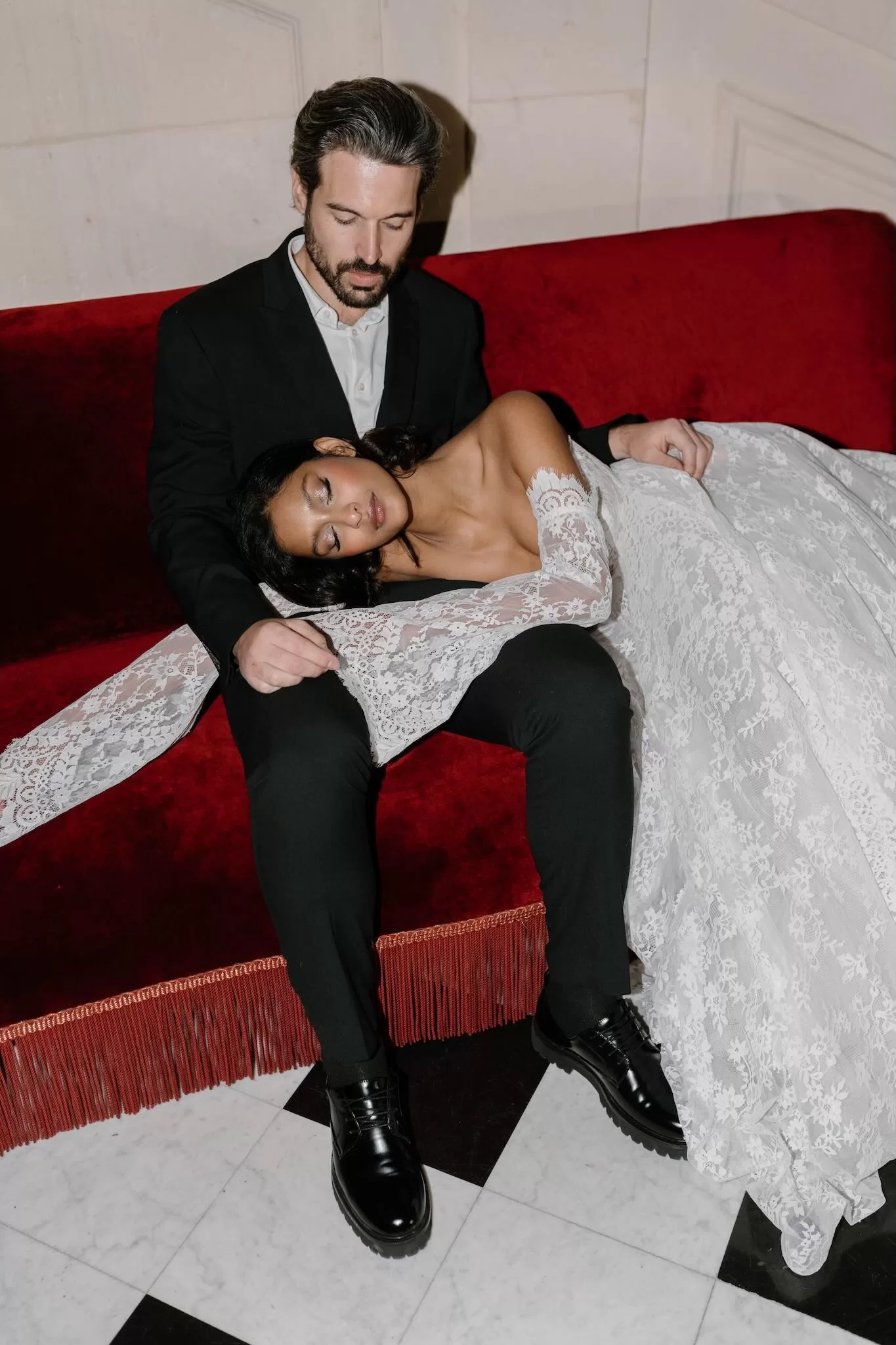 Bride rests peacefully in groom’s lap on a deep red velvet sofa, her lace gown draped around them, by Bahr Studios.