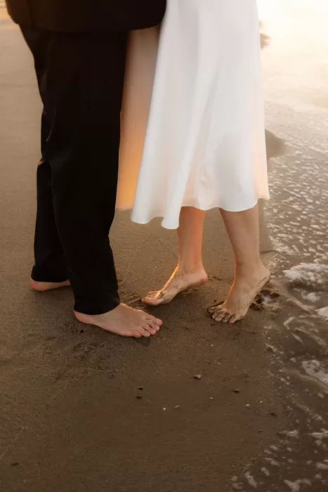 A barefoot couple stands close together on wet sand, the golden sunset casting a warm glow on their feet.