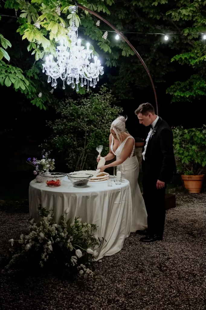 Bride and groom decorate their wedding cake under a chandelier in the garden at Borgo Sant'Ambrogio.