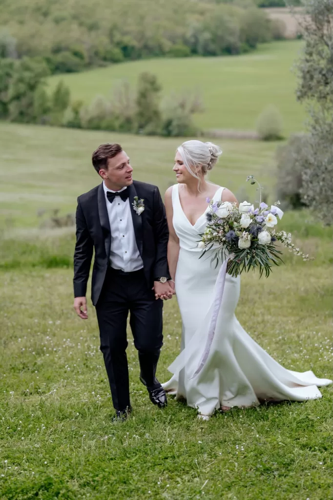 Bride and groom walking hand-in-hand through a Tuscan meadow at Borgo Sant'Ambrogio.