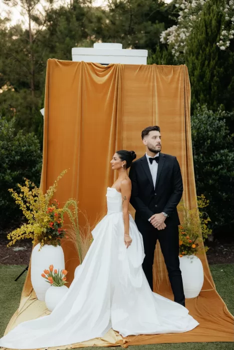 Bride and groom strike confident pose in front of mustard velvet ceremony backdrop at The Bradford NC.