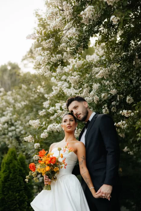 Couple stands beneath white flowering branches holding a bold orange bouquet at The Bradford NC.