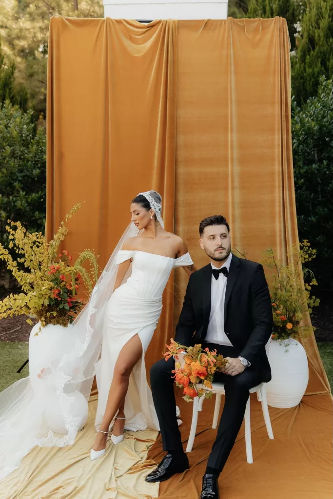 Bride in off-shoulder gown poses beside groom against a velvet backdrop at The Bradford NC.