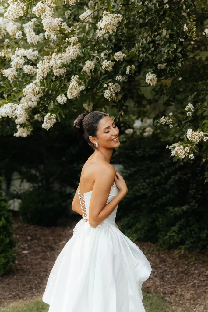 Bride smiles joyfully in lace-back strapless gown under flowering branches at The Bradford NC.