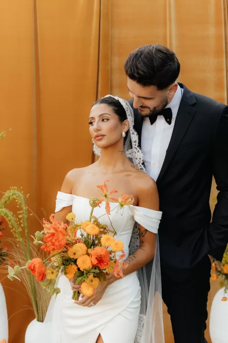 Groom leans in toward bride holding orange bouquet in front of velvet backdrop at The Bradford NC.