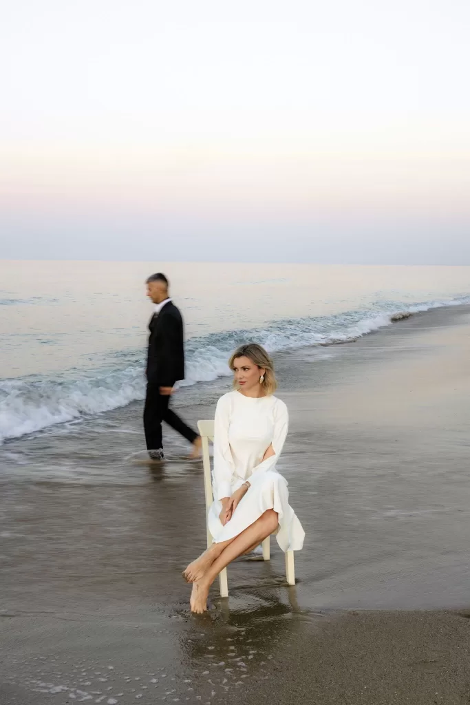 The bride sits poised at the shoreline as the groom walks away, a moment of distance and reflection.