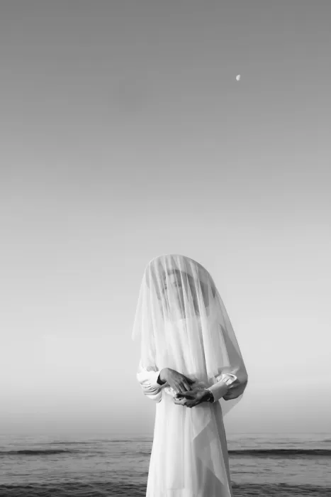 A serene bride stands veiled by the ocean under the pale moon, captured in still black and white.