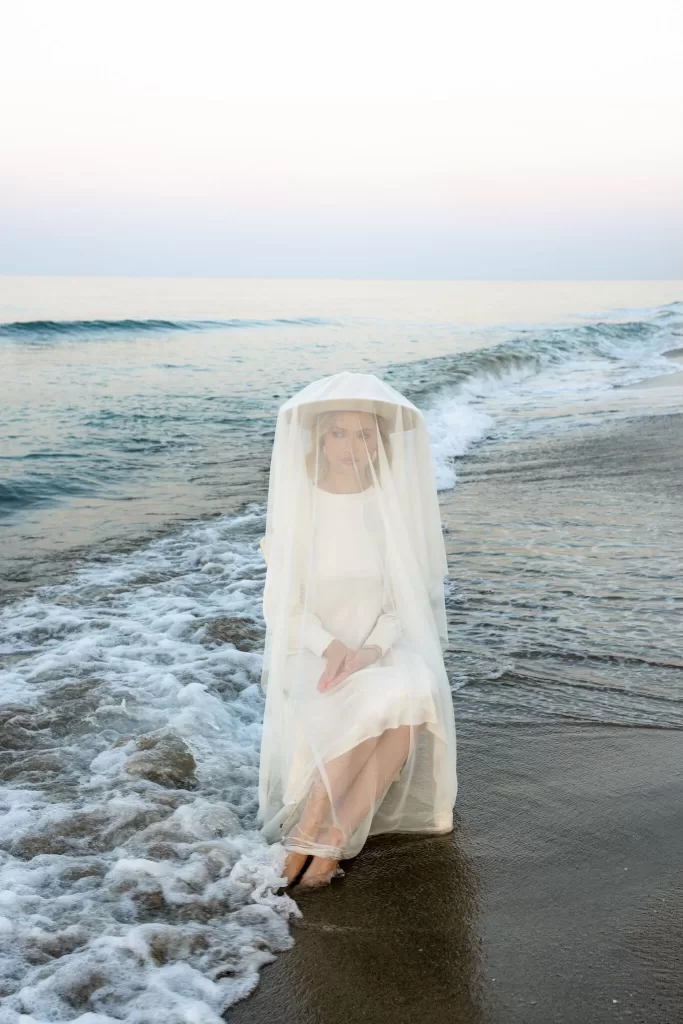 The bride sits on the shore with the tide at her feet, her translucent veil draped over her like a shell.