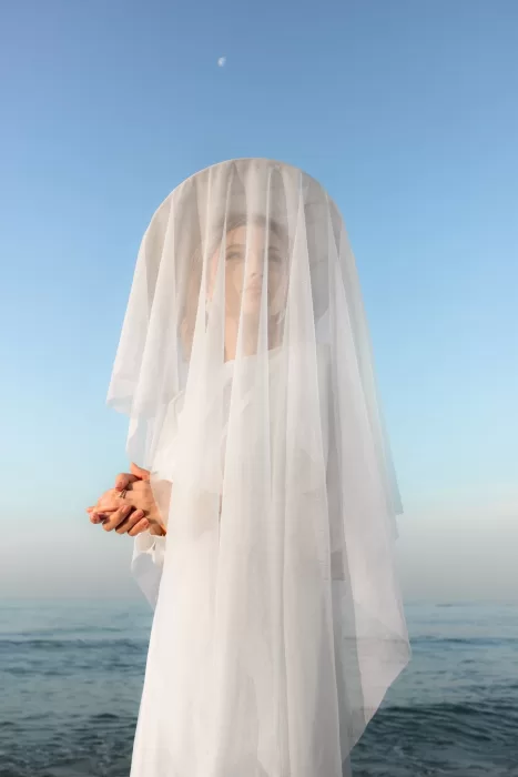 A bride beneath a wide veil gazes forward with the moon above and the ocean stretching behind her.