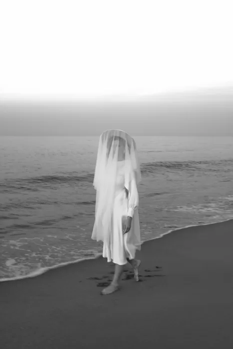 The veiled bride walks barefoot along the shore, her figure ghostlike in the soft evening haze.