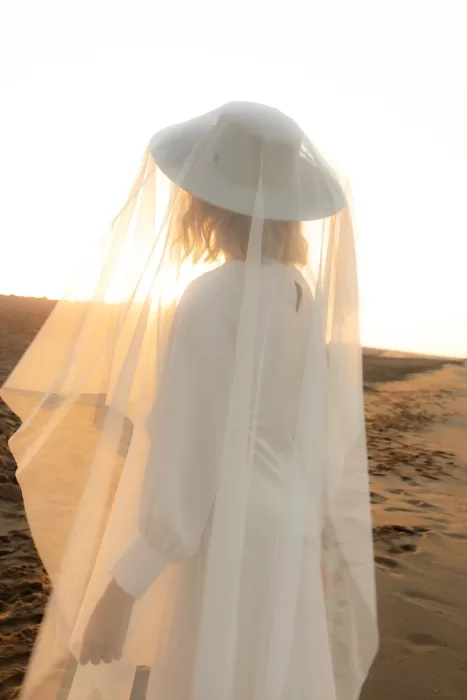 The bride walks alone at golden hour, her wide-brimmed veil glowing in the last light of day.