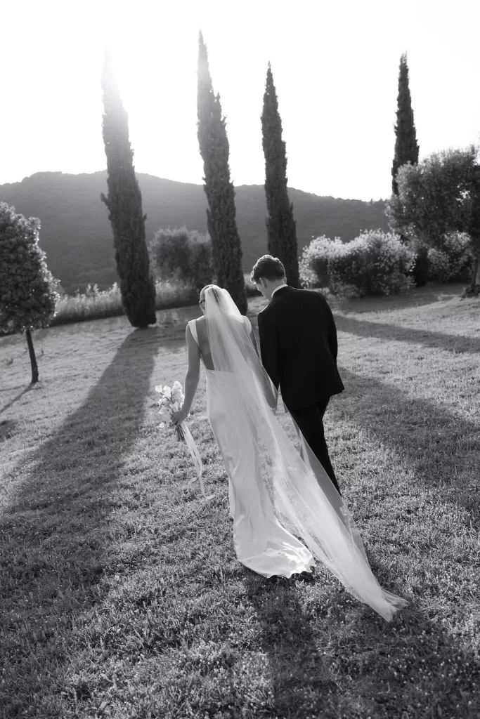 Bride and groom walk hand in hand through the gardens of Casa Olivi, framed by tall cypress trees in soft sunset light.