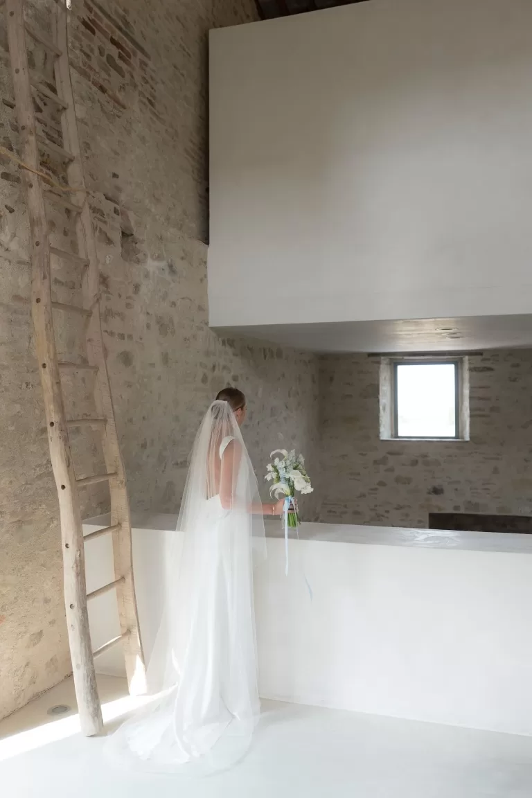 Bride stands quietly in Casa Olivi’s minimalist stone interior, holding her bouquet and gazing out the window.