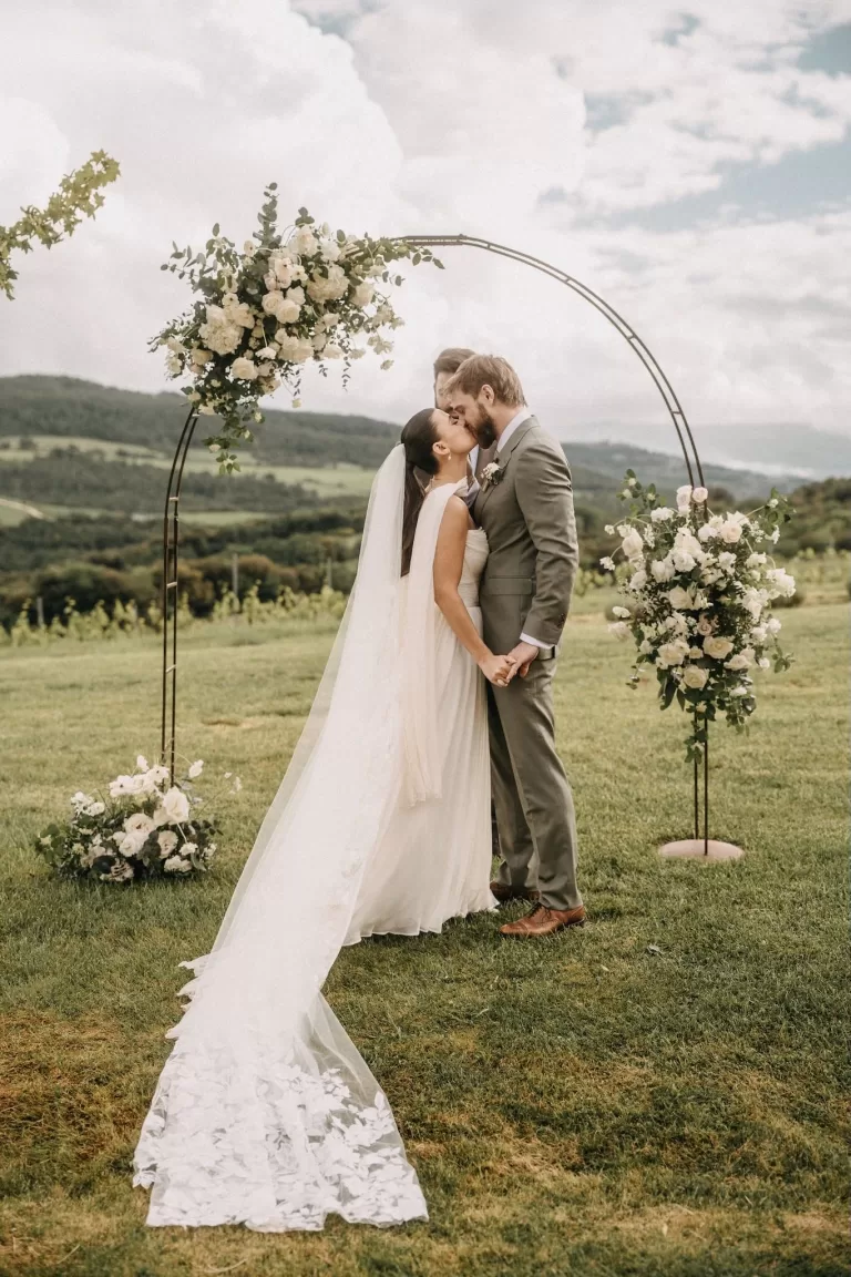 Bride and groom share a kiss under a floral arch on the lush ceremony lawn at Castello di Casole.