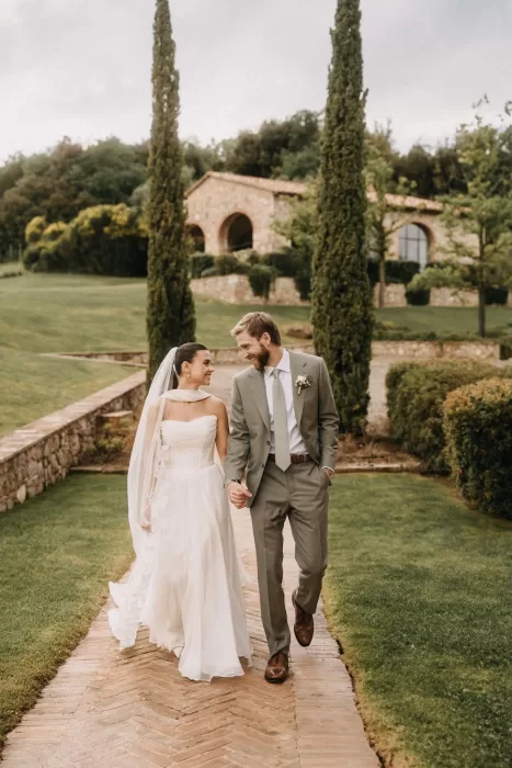 Just-married couple walk hand in hand through the manicured grounds of Castello di Casole, smiling warmly.