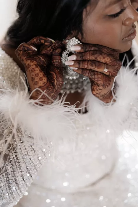 Close-up of the bride adjusting her sparkling earrings, showcasing intricate henna and a feather-trimmed beaded gown.