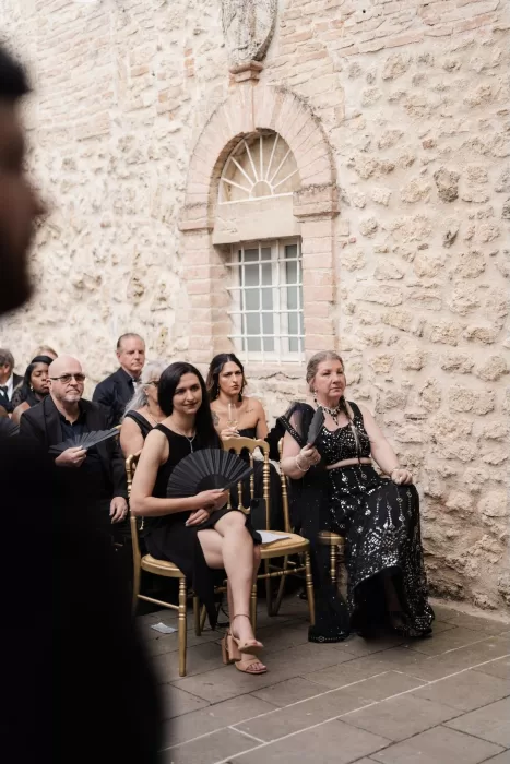 Wedding guests dressed in black seated at the Castello di Montignano ceremony, fanning themselves in the summer heat.