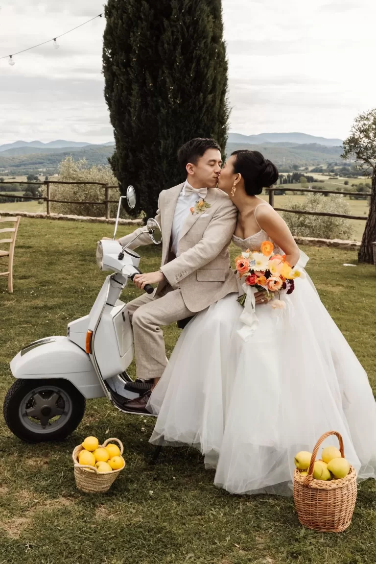 Bride and groom sharing a kiss on a vintage Vespa with colourful bouquet and Tuscan hills at Conti di San Bonifacio.