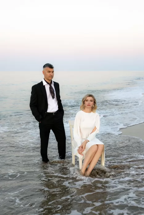 The bride sits on a white chair in the surf while the groom stands beside her, both framed by gentle evening waves.