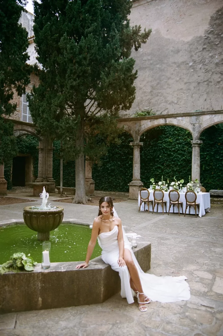 Bride seated by the courtyard fountain at Finca Es Convent, with reception tables set in the background.