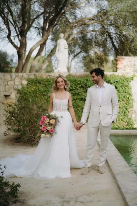 The bride and groom walk hand in hand beside a peaceful courtyard fountain at Finca Son Collell, smiling at each other.