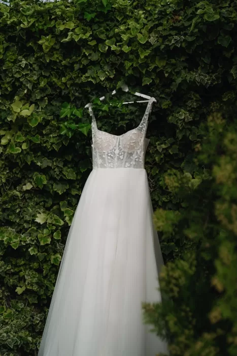 A romantic wedding gown with floral bodice and flowing tulle skirt hanging against lush green ivy at Finca Son Collell.