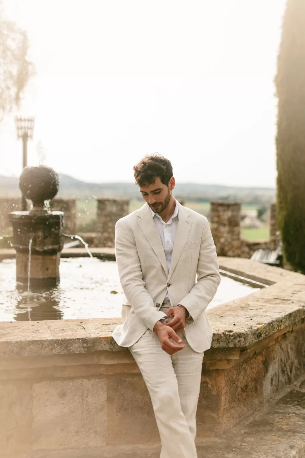 Groom in light linen suit by the stone fountain with views of the countryside at Finca Son Collell