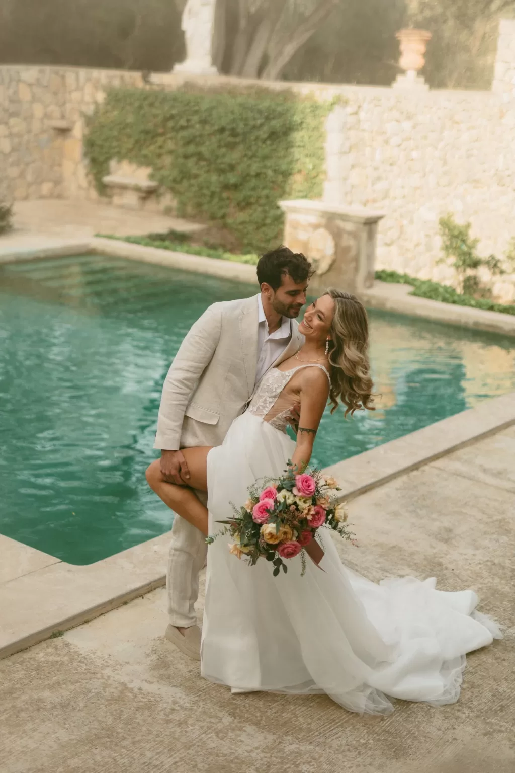 Romantic moment by the pool as the groom lifts the bride holding a vibrant bouquet at Finca Son Collell